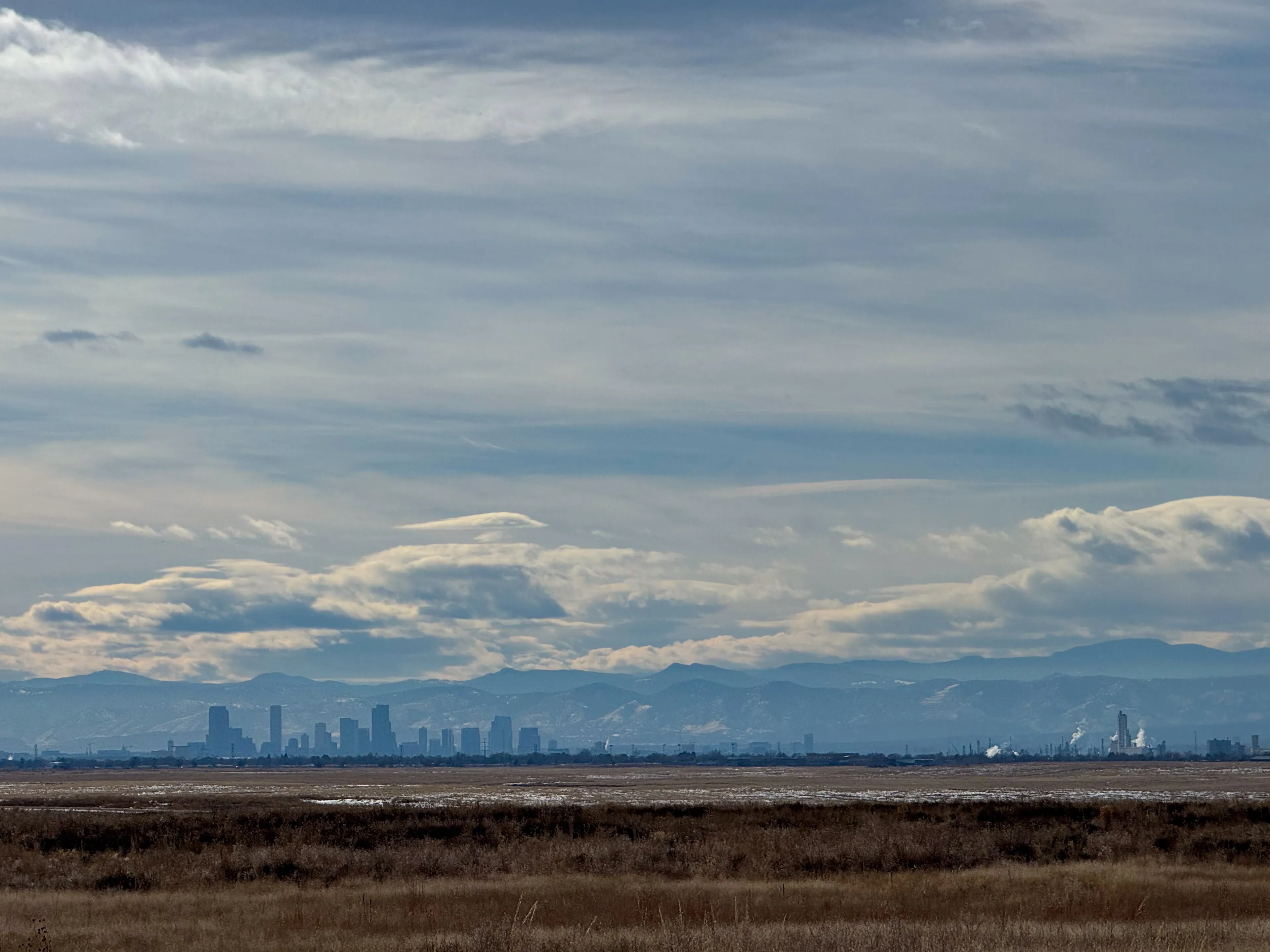 Denver skyline under a winter sky with the snow-covered Front Range behind, seen from the eastern plains